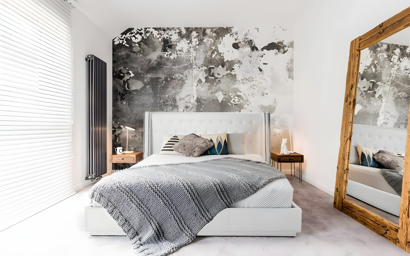 Une chambre claire avec un lit rembourré blanc à tête capitonnée, un jeté gris et un grand miroir au cadre de bois
